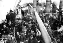 A group of people gathered on the crowded deck of a ship, with smokestacks and rigging visible in the background.