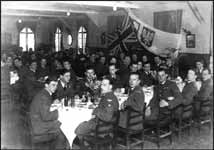 A large group of men in military uniforms sit at long tables in a dining hall with a British flag and crest displayed on the back wall.