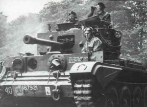 Black and white photo of three soldiers riding in a British Crusader tank, with trees visible in the background.