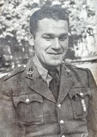 Black-and-white photo of a man in a military uniform, outdoors, facing the camera and smiling, with trees blurred in the background.