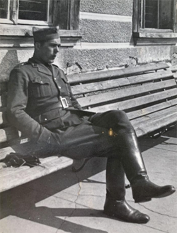 A man in a military uniform sits on a wooden bench outdoors, with his legs crossed and hands resting on his lap.