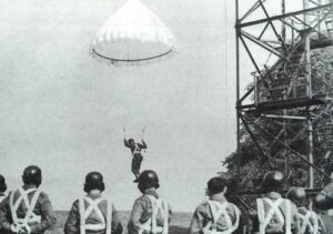 A parachutist descends during training as a group of soldiers in helmets and harnesses observe near a metal tower structure.