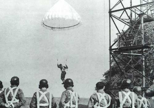 A parachutist descends during training as a group of soldiers in helmets and harnesses observe near a metal tower structure.