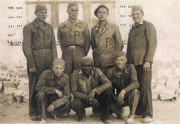 Seven prisoners in marked uniforms pose in front of a brick wall; three are kneeling in front, four stand behind, with neutral expressions.