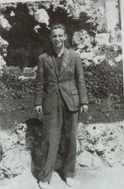 A young man in a suit stands outdoors in front of a rocky wall, smiling at the camera.