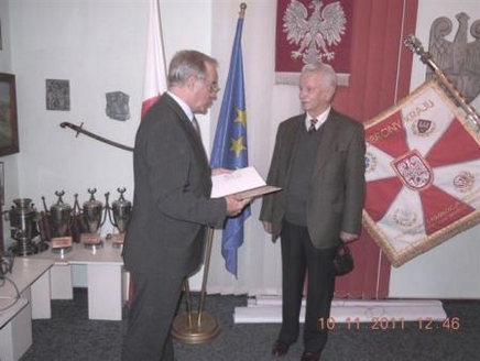 Two men in suits stand in an office with flags, trophies, and a banner. One man holds a document, whilst the other holds a hat. Date stamp on the photo reads "10/11/2011 12:46".