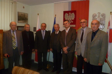 Seven men in suits stand in a row indoors, in front of Polish flags and an emblem, posing for a group photo.