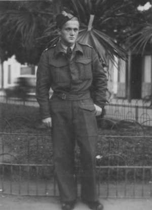 A man in a military uniform stands in front of a fence and a large plant, looking slightly to the side. Black and white photo.