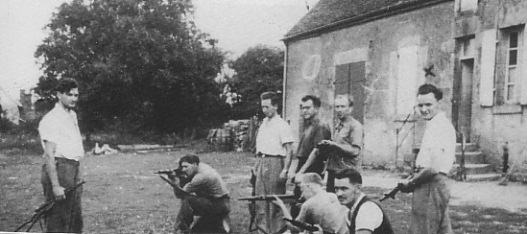 A group of men, some kneeling and some standing, aim rifles outside a stone building in a rural setting.
