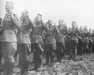 A line of uniformed soldiers stands outdoors, facing forwards with their right hands raised in salute.