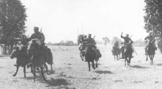 A group of soldiers on horseback charge across an open field, with trees visible in the background.