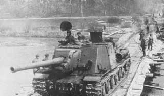 Black and white photo of a military tank with soldiers on top crossing a bridge, with other soldiers walking alongside on a wooden walkway.