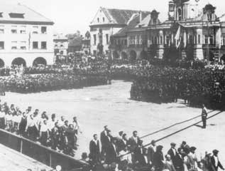 Large crowd gathers in a town square with people marching in rows and historic buildings in the background.