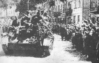 Crowds line both sides of a street as a military tank, covered with soldiers and foliage, drives through a city during wartime.