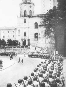 Soldiers stand in formation on a city street near a large building with a tower, whilst a few civilians walk nearby.