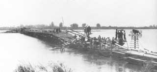A group of people work together building a makeshift wooden bridge across a river, with trees visible in the background.