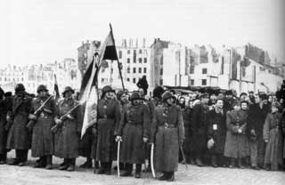 A group of soldiers stand in uniform holding flags and swords, with ruined buildings and a crowd of civilians in the background.