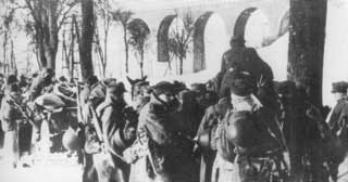 A group of soldiers in winter clothing gather outdoors near an arched stone structure, with snow-covered ground and bare trees visible.