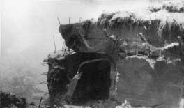 A partially destroyed concrete pillbox with exposed reinforcing bars and earth covering, surrounded by rubble and fog.
