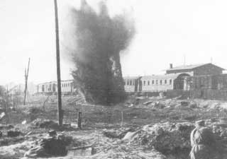 A large explosion occurs near a building complex, with debris and smoke rising into the air; the ground is rough and a few people are visible in the foreground.