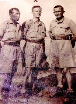 Three men in military-style shirts and shorts pose outdoors in a sepia-toned photograph.