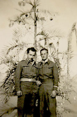 Two men in military uniforms stand in front of a decorated palm tree. The photo is in black and white and appears to be from a past era.