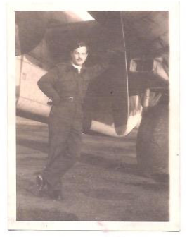 A man in military uniform stands, leaning against the propeller of a large aeroplane in a black-and-white photograph.