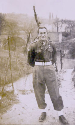 A man in a military uniform stands outdoors on a dirt path near a stream, smiling at the camera with trees and a bridge in the background.
