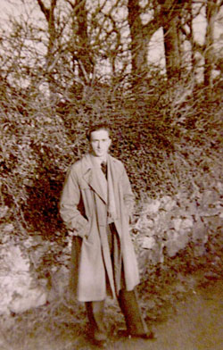 A man in a long coat stands outdoors in front of a stone wall and dense bushes, with bare trees in the background.
