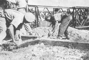 Two workers bend over while mixing or spreading material at a construction site with a metal bridge framework in the background.