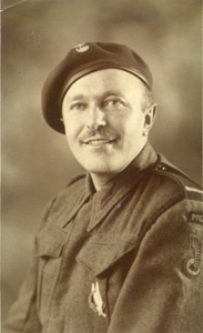 A man in military uniform and beret poses for a portrait, smiling slightly. The uniform features insignia and badges on the shoulder and chest.