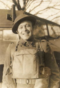 A smiling soldier in uniform and helmet stands in front of a military vehicle on a sunny day.