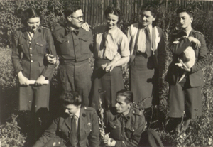 Seven people, some in military uniforms, pose outdoors in a garden; one person on the right holds a cat.