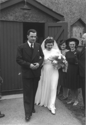 A bride in a white dress and veil holding flowers walks beside a man in a suit, exiting a building, with several people standing and smiling nearby.