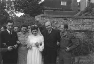 A bride in a white dress and veil stands with four people—two men in suits, a woman in a dress, and a man in military uniform—outside near a brick wall and house.