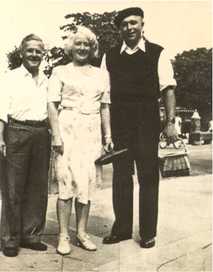 Three adults pose outdoors on a sunny day; two men stand on either side of a woman, all wearing mid-20th-century attire. Trees and a park setting are visible in the background.