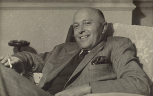 A man in a suit sits on a patterned sofa, smiling and holding a cigarette. The background shows a textured wall and part of a mantelpiece.