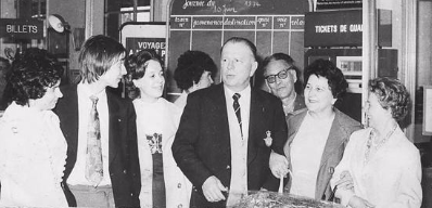 A group of seven adults stand together in a railway or ticket station, talking and smiling. Signs in French are visible in the background.