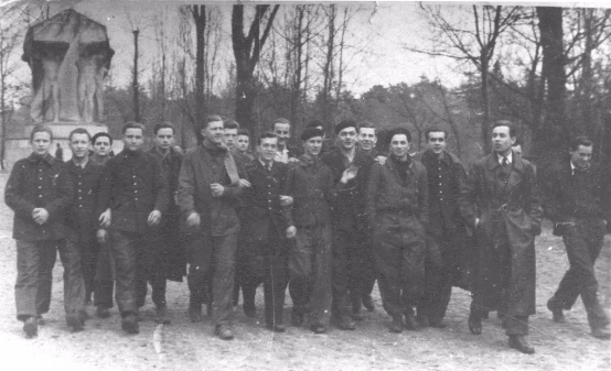 A group of men in coats and uniforms stand outdoors in front of leafless trees and a covered statue or monument, posing for a black and white photo.