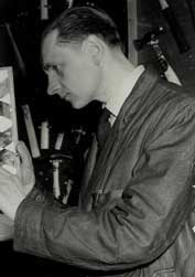 A man in a suit jacket examines an object closely, viewed in profile, with various tools or instruments visible in the background.