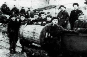 A group of miners in work clothes and hats pose around a mine trolley outside an industrial building.