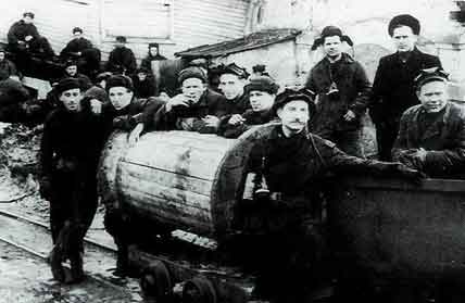 A group of miners in work clothes and hats pose around a mine trolley outside an industrial building.