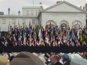 A large group of people in formal attire hold colourful flags on a stage in front of a historic building, whilst a crowd watches in the foreground.
