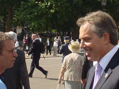 Two men converse outdoors in a crowd; one is wearing a suit and tie, the other is casually dressed. People walk in the background near trees.