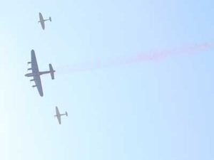 Three vintage military aeroplanes fly in formation in a clear sky, with the largest plane emitting a faint red trail behind it.