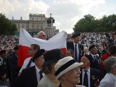 A large crowd of people, many wearing hats and medals, gathers outdoors. One person holds a large red and white flag. A historic building is visible in the background.