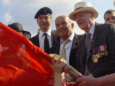 A group of men, one in a military beret and another wearing medals and a white hat, stand together as a child waves a large red flag in the foreground.