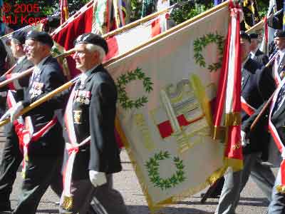 A group of uniformed men march in a parade, carrying flags and banners decorated with red, white, and green colours.