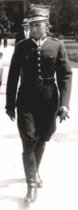 A man in a dark military uniform with a peaked cap walks outdoors on a paved path, with another person visible in the background.