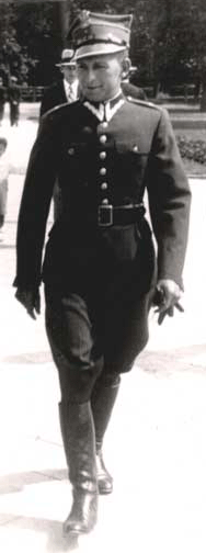 A man in a dark military uniform with a peaked cap walks outdoors on a paved path, with another person visible in the background.